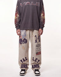 A person wearing a dark gray long-sleeve sweatshirt with 'VALE' across the chest and flame graphics on the sleeves, paired with beige sweatpants featuring purple collegiate-style lettering and graphics, and black-and-white sneakers, against a plain white studio background.