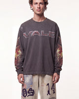 A man wearing a dark gray long-sleeve t-shirt with 'VALE' printed across the chest in metallic letters and flame graphics on the sleeves, paired with beige pants featuring graphic prints.