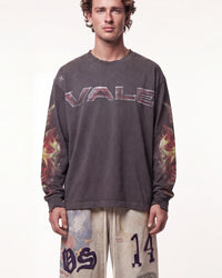 A man wearing a dark gray long-sleeve t-shirt with 'VALE' printed across the chest in metallic letters and flame graphics on the sleeves, paired with beige pants featuring graphic prints.