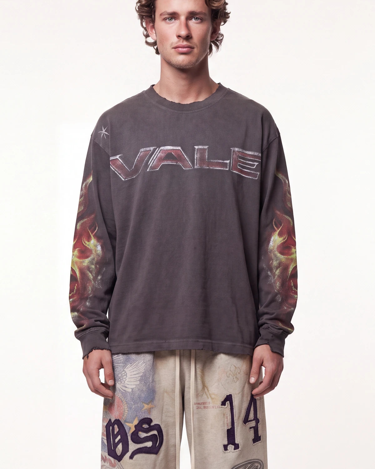 A man wearing a dark gray long-sleeve t-shirt with 'VALE' printed across the chest in metallic letters and flame graphics on the sleeves, paired with beige pants featuring graphic prints.