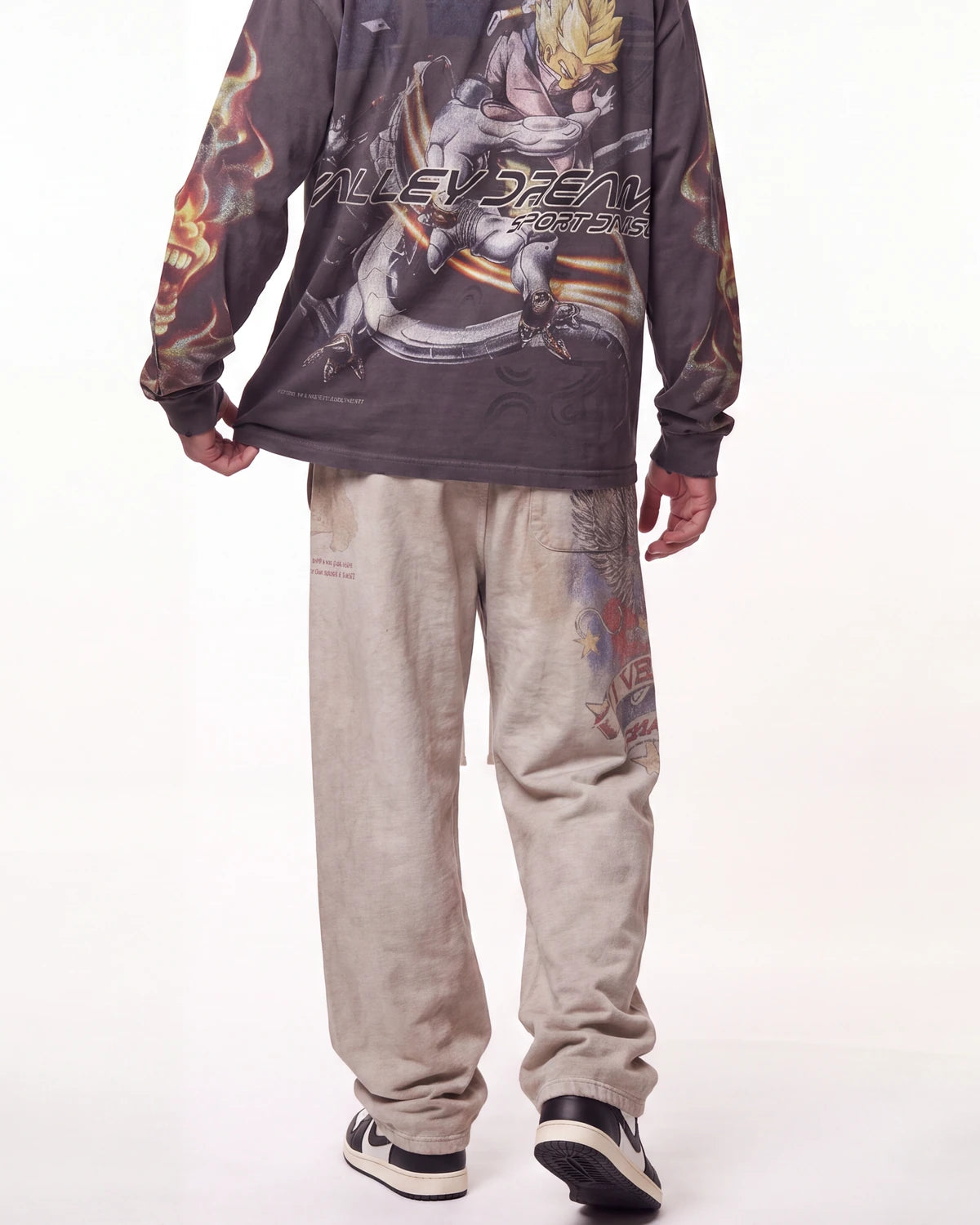 A person shown from the back wearing a dark gray long-sleeve graphic shirt with anime artwork and the text 'NILEYSEAN BEASTS' across the back, paired with light beige sweatpants and black-and-white sneakers, against a plain white studio background.