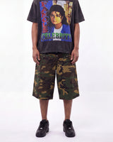 A model wearing a faded black graphic t-shirt with a colorful 'Famous Celebrity' Michael Jackson print, paired with green and brown camouflage shorts and black sneakers, against a plain white background.