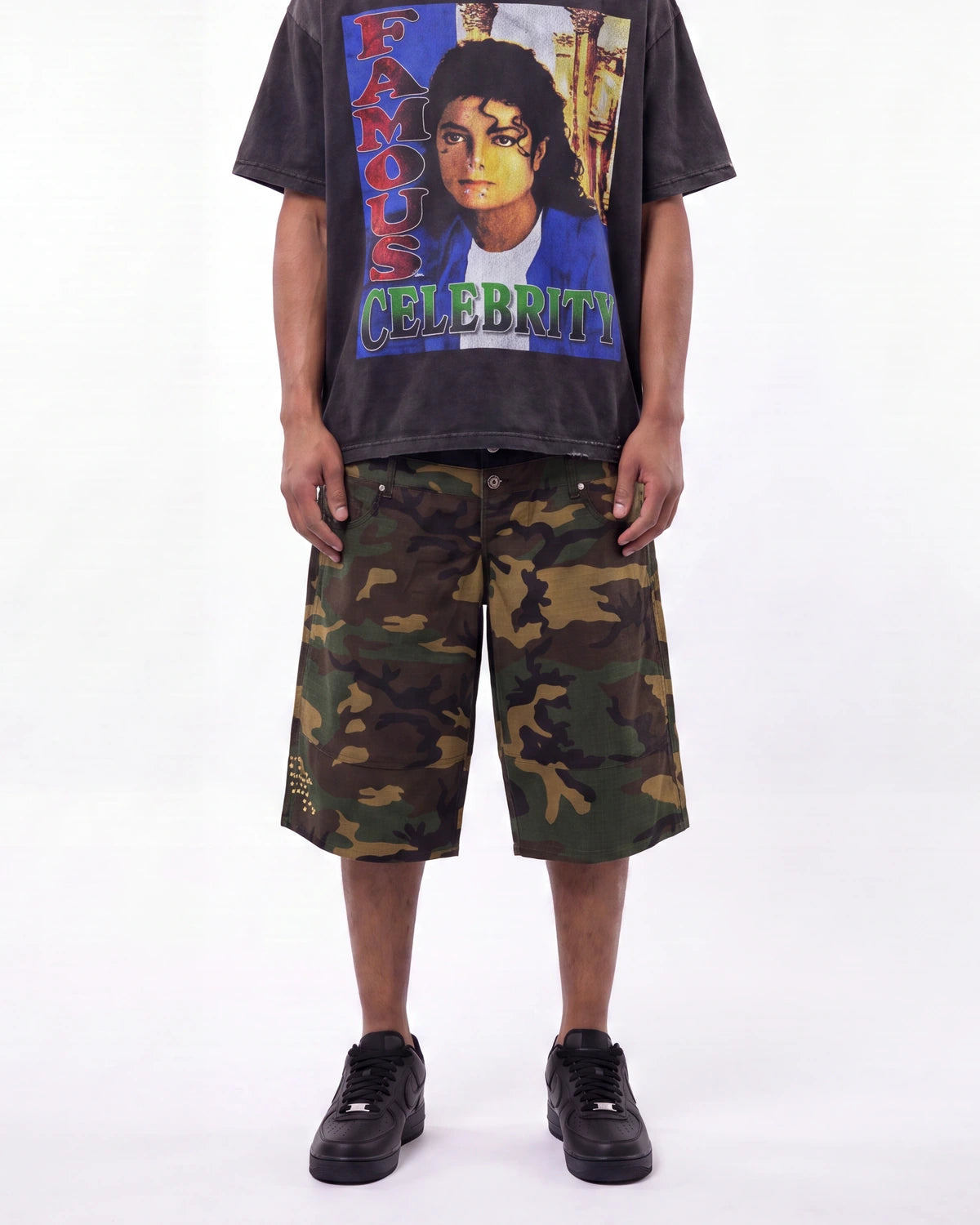 A model wearing a faded black graphic t-shirt with a colorful 'Famous Celebrity' Michael Jackson print, paired with green and brown camouflage shorts and black sneakers, against a plain white background.