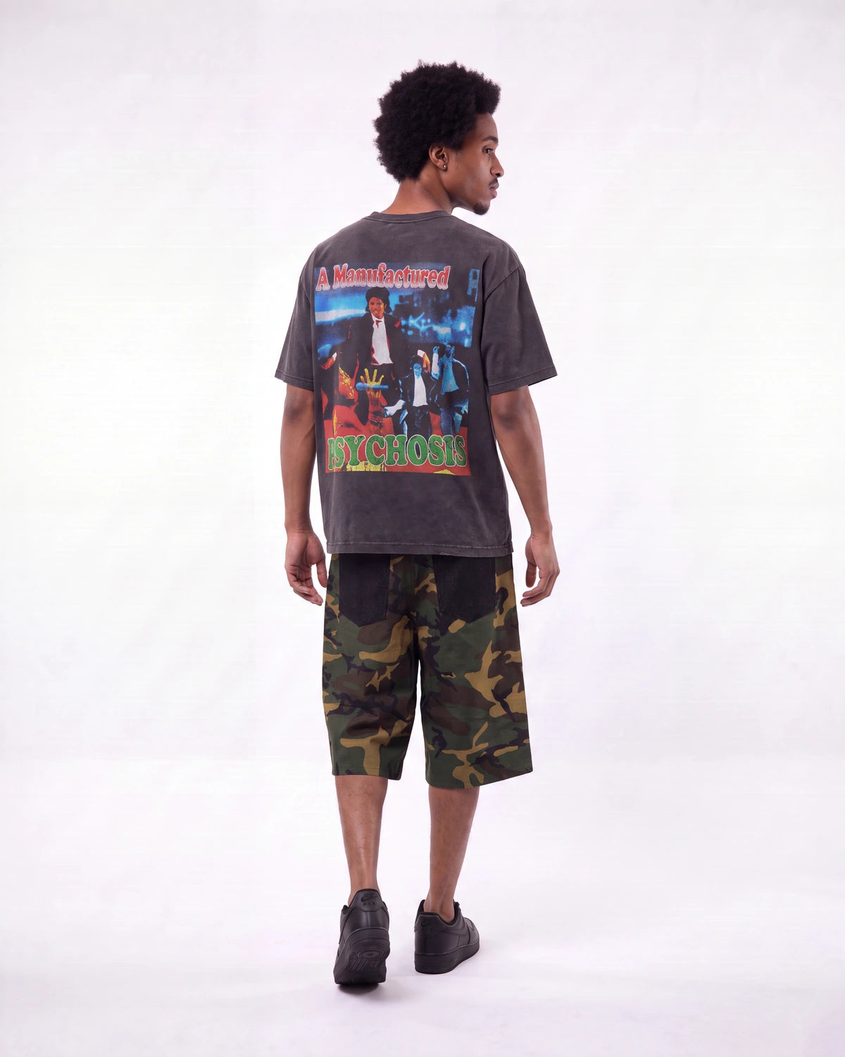 A man shown from the back wearing a dark gray graphic t-shirt with a large 'Psycho' print, paired with green and brown camouflage shorts, against a plain white studio background.