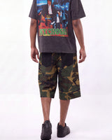 A person shown from the waist down wearing a dark graphic t-shirt and green camouflage knee-length shorts, standing against a plain white studio background.
