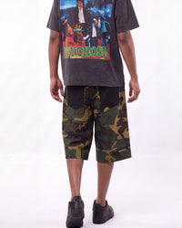 A person shown from the waist down wearing a dark graphic t-shirt and green camouflage knee-length shorts, standing against a plain white studio background.