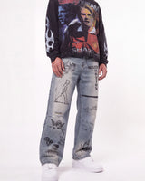 A person wearing a black long-sleeve graphic sweatshirt with football player prints, light-wash distressed jeans covered in graffiti-style drawings and text, and white sneakers, photographed against a plain white studio background.