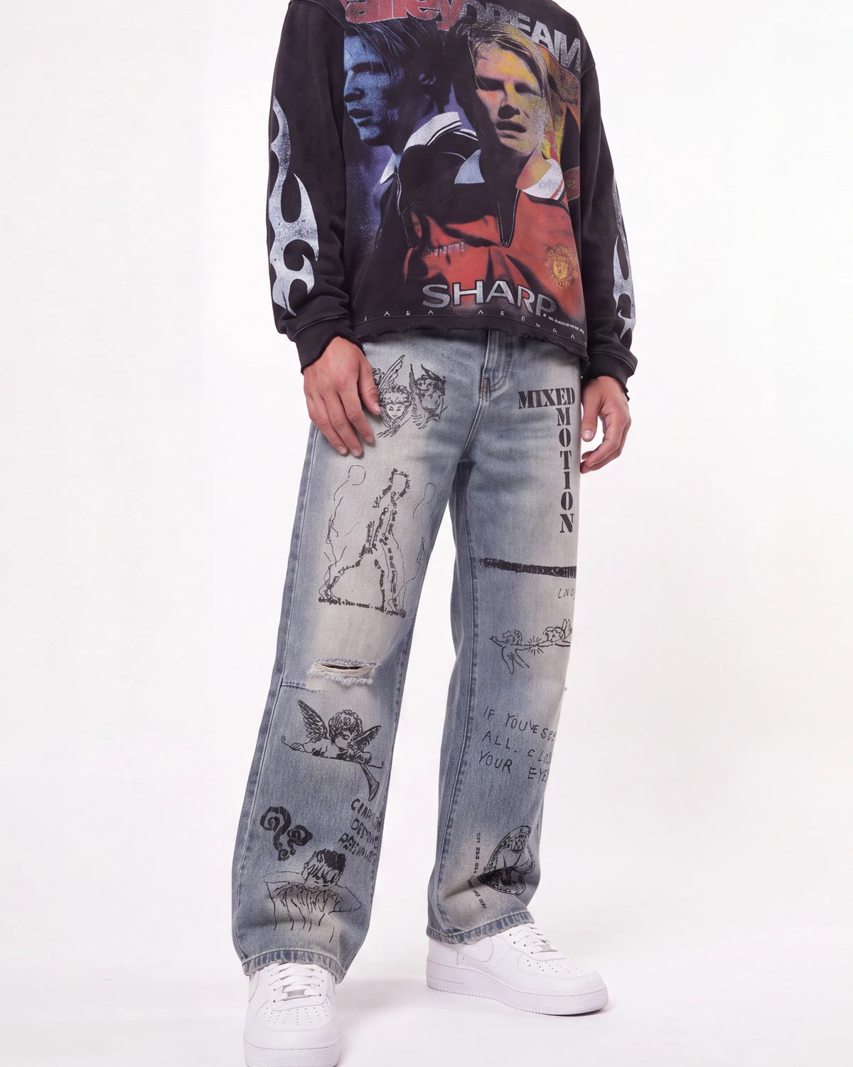 A person wearing a black long-sleeve graphic sweatshirt with football player prints, light-wash distressed jeans covered in graffiti-style drawings and text, and white sneakers, photographed against a plain white studio background.