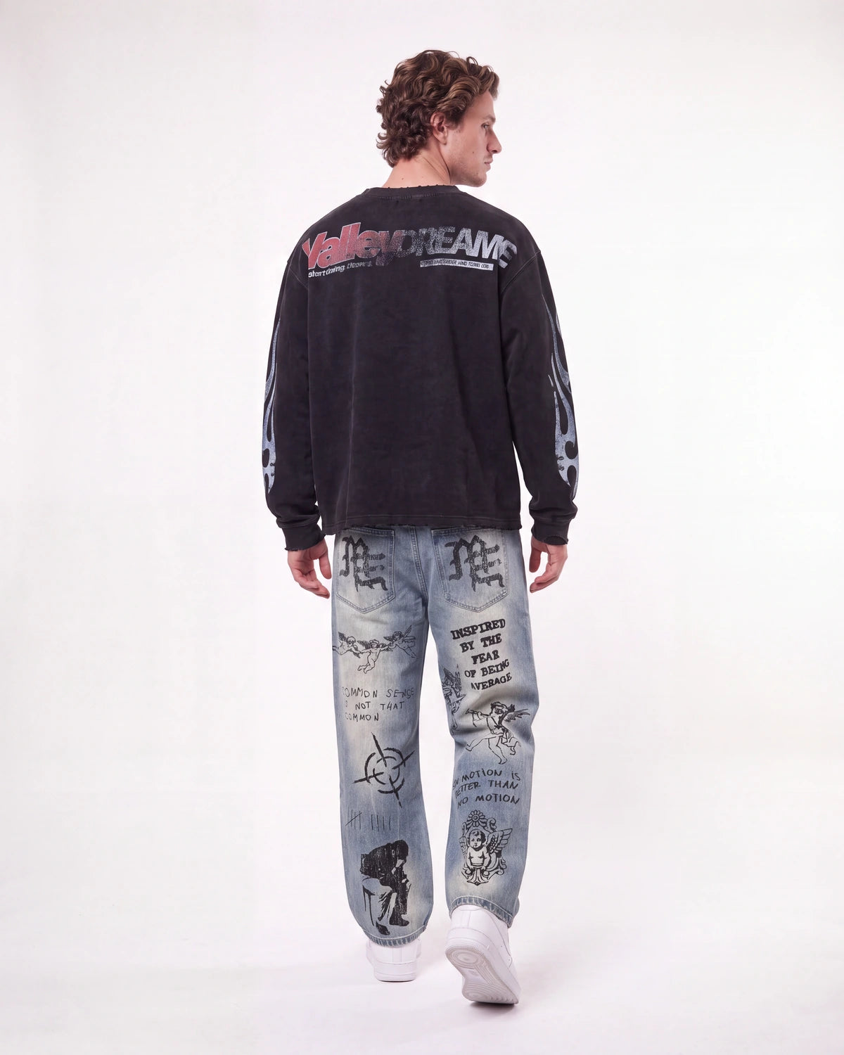 A man shown from the back wearing a black long-sleeve sweatshirt with 'Valley Dream' text on the back and light blue graphic jeans, against a plain white studio background.