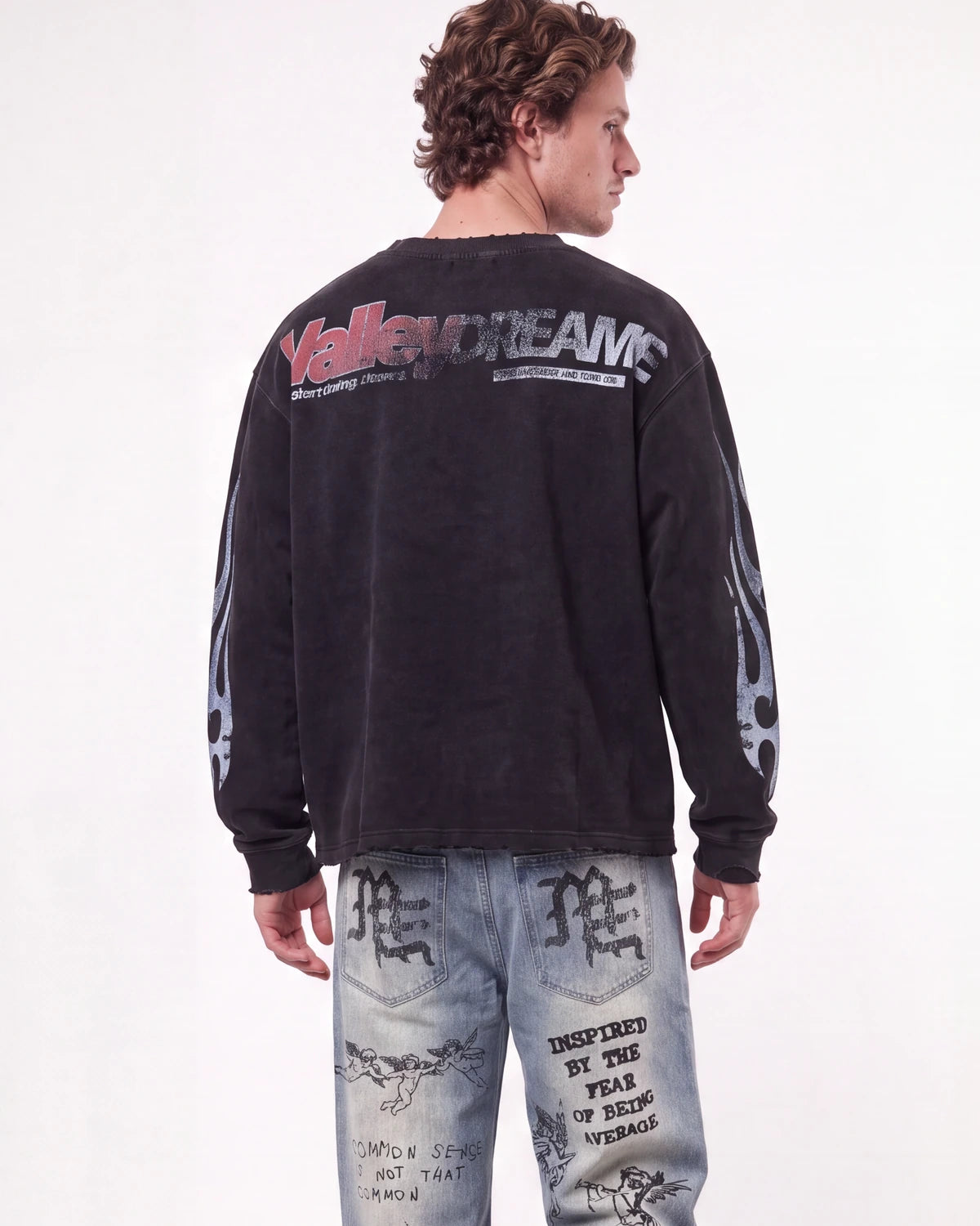 A man shown from the back wearing a faded black long-sleeve graphic sweatshirt with 'VallenDreams' printed across the upper back and distressed light-wash denim jeans with graphic prints.