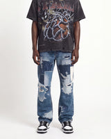 A model wearing a faded black graphic t-shirt, distressed blue denim jeans with ripped details, and black and white sneakers, standing against a plain white studio background.