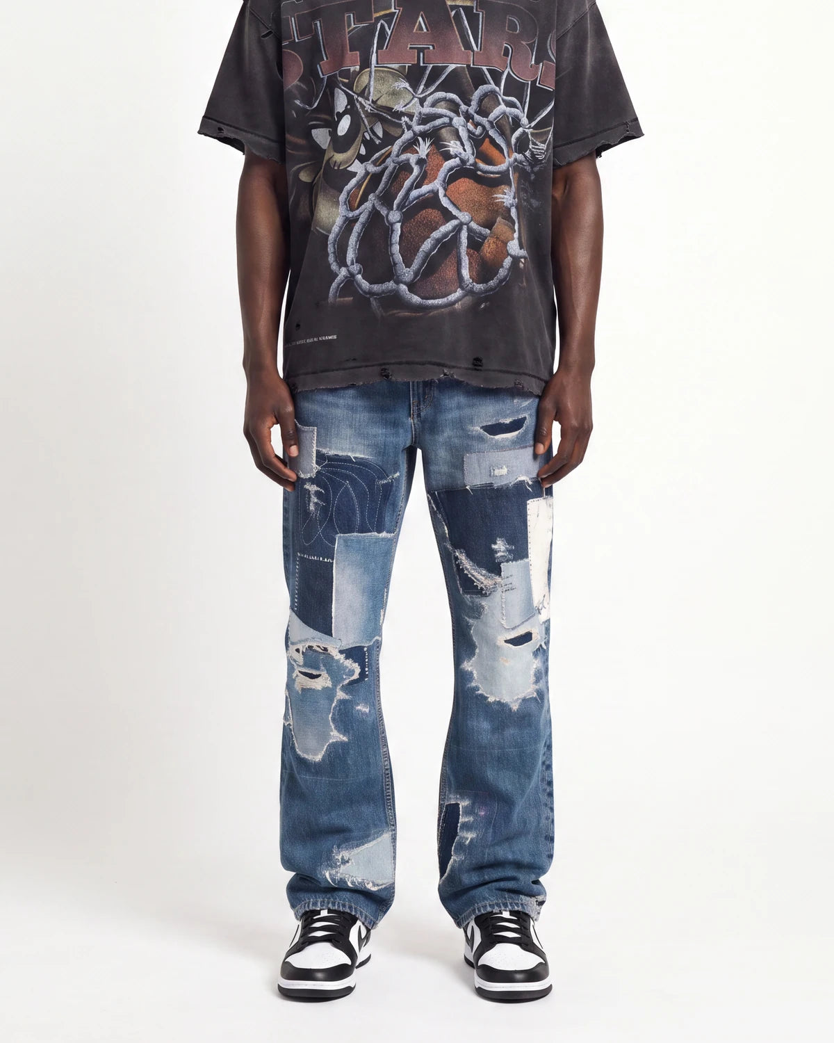 A model wearing a faded black graphic t-shirt, distressed blue denim jeans with ripped details, and black and white sneakers, standing against a plain white studio background.