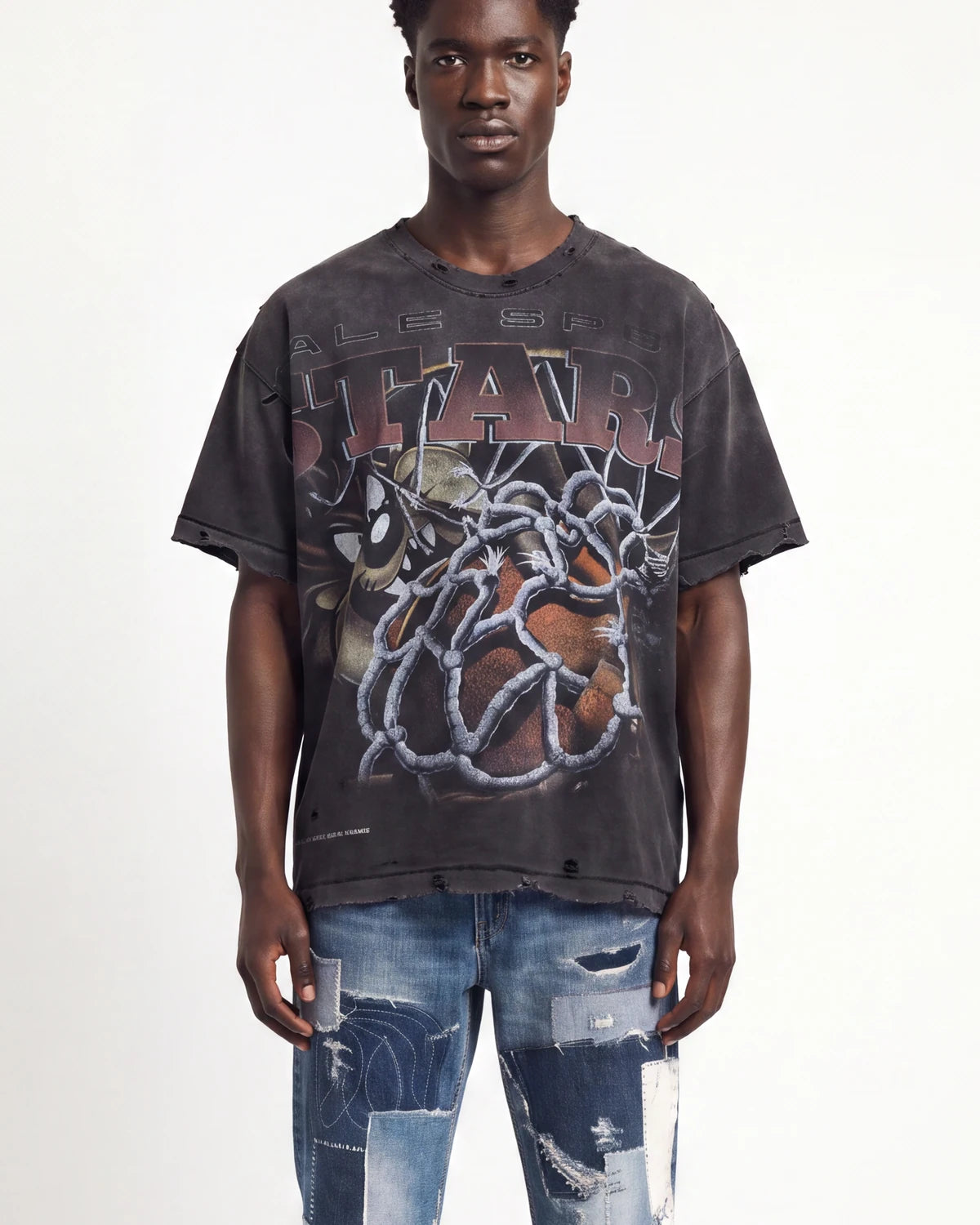 A man wearing an oversized faded black graphic t-shirt with distressed details and a large front print, paired with ripped blue jeans, against a plain white background.
