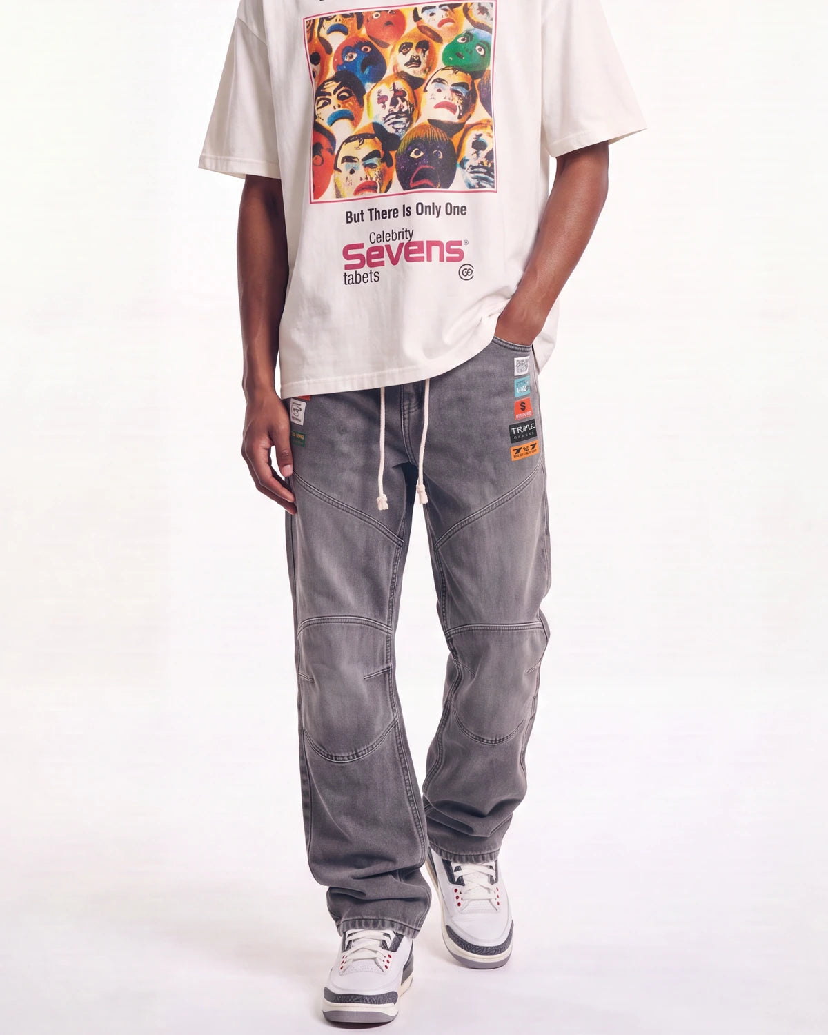 A person wearing a cream graphic T-shirt and faded gray baggy sweatpants with white sneakers, standing against a white studio background.