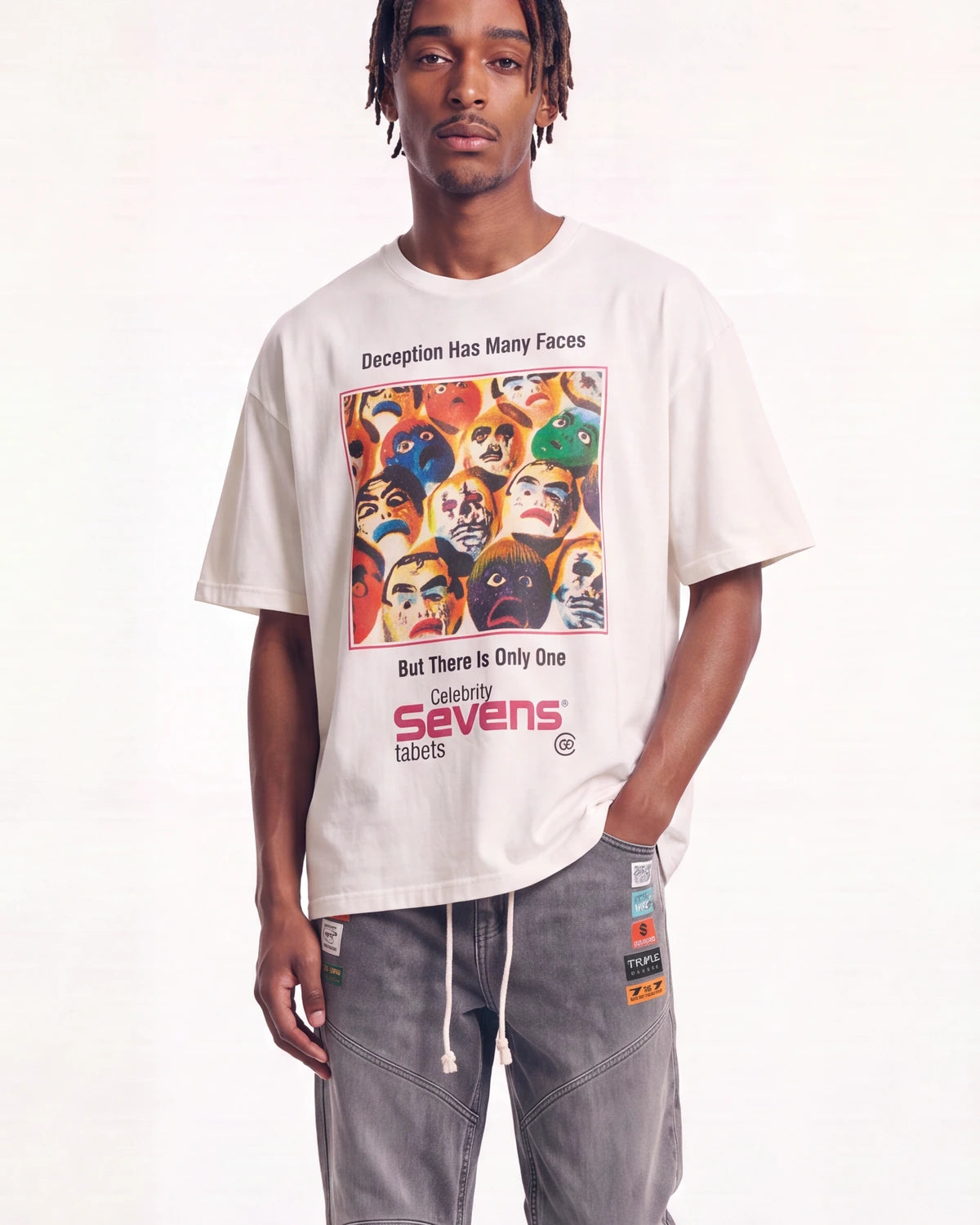 A man wearing an oversized off-white t-shirt with a colorful graphic print of faces and the text 'Deception Has Many Faces But There Is Only One Galaxy Sevens labels', paired with gray drawstring pants, against a plain white background.