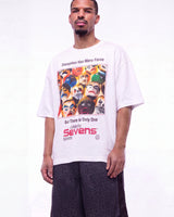 A man wearing a white graphic t-shirt with a colorful print of Lucha Libre masks and text, paired with dark patterned shorts, against a plain white studio background.