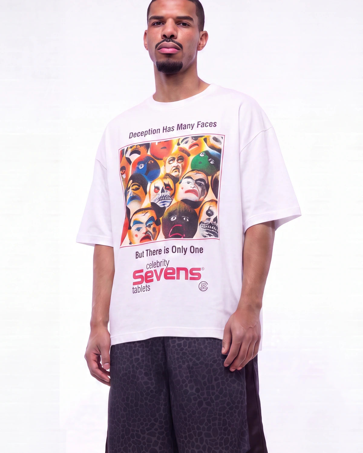 A man wearing a white graphic t-shirt with a colorful print of Lucha Libre masks and text, paired with dark patterned shorts, against a plain white studio background.