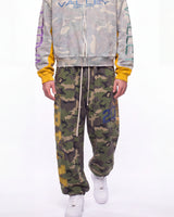 A person wearing a light gray camouflage zip-up hoodie with yellow and purple sleeve accents, green camouflage jogger pants with a blue graphic, and white sneakers, photographed against a plain white studio background.