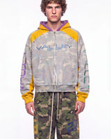 A male model wearing a distressed zip-up hoodie with a light gray camouflage pattern, yellow sleeves and hood lining, and purple interior hood lining. The hoodie features 'VALLEY SPORT' printed across the chest in blue.