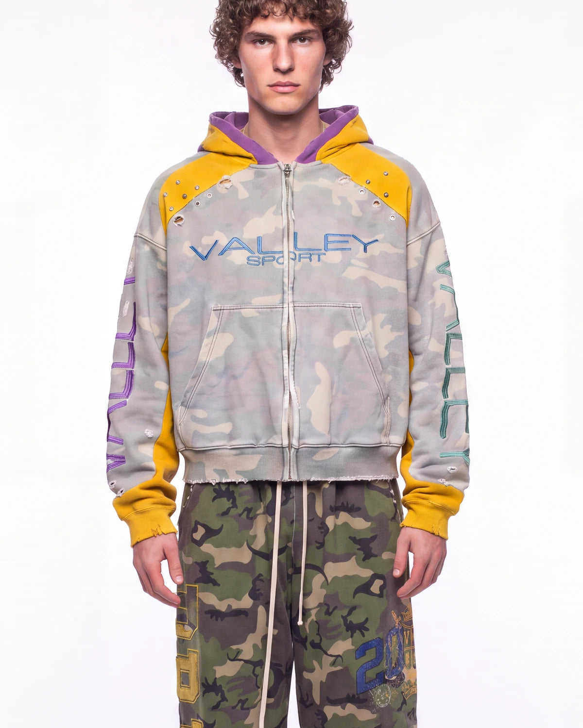 A male model wearing a distressed zip-up hoodie with a light gray camouflage pattern, yellow sleeves and hood lining, and purple interior hood lining. The hoodie features 'VALLEY SPORT' printed across the chest in blue.
