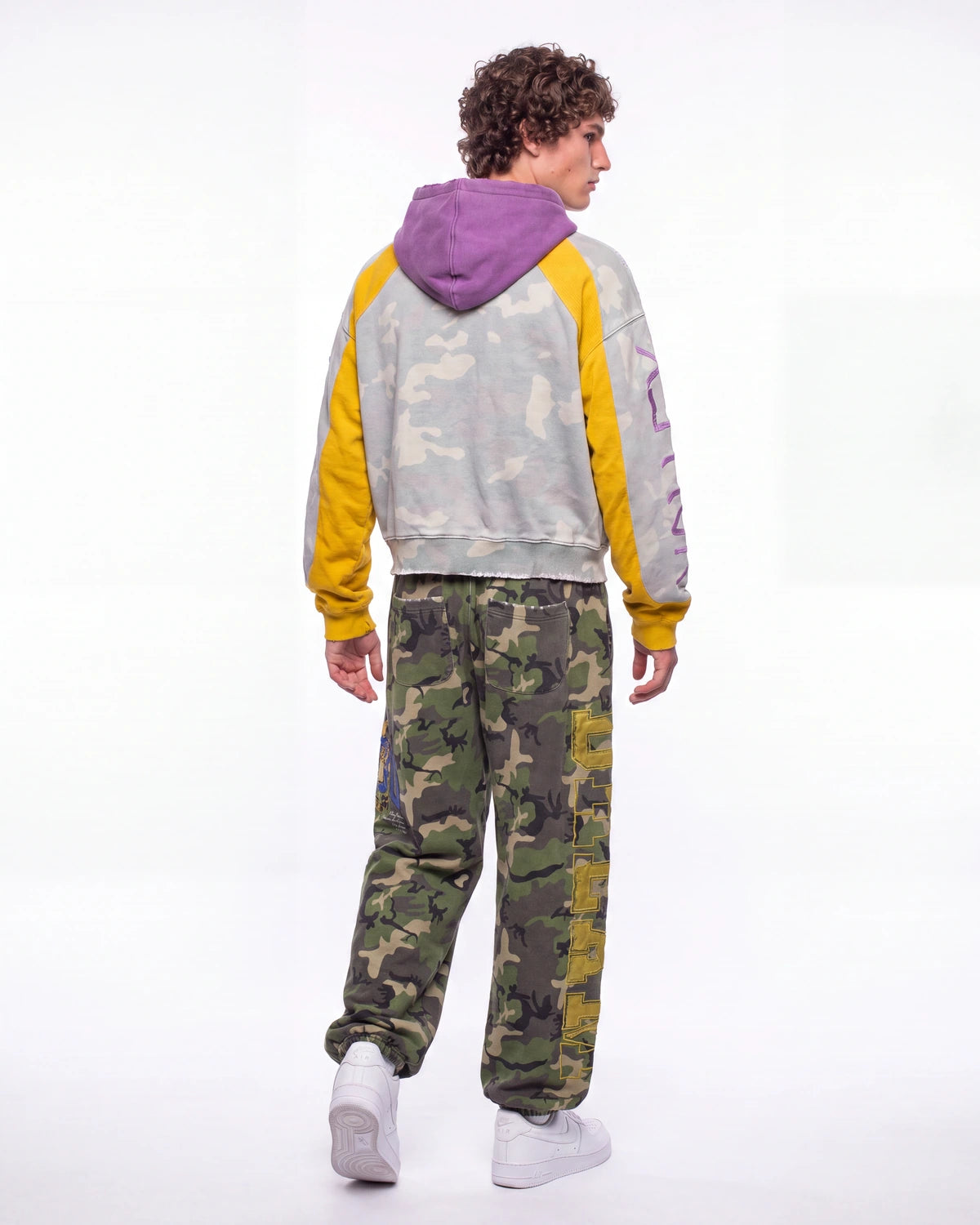 A model shown from the back wearing a gray and yellow color-block hoodie with a purple hood, paired with green camouflage sweatpants and white sneakers, against a plain white studio background.