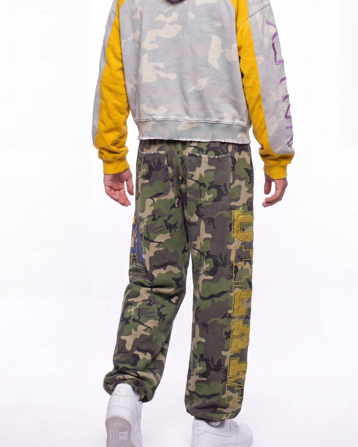 A person shown from the back wearing camouflage cargo sweatpants with yellow side panels, a gray and yellow camouflage hoodie, and white sneakers against a plain white background.