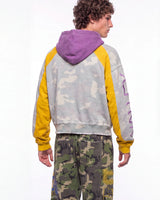A model shown from the back wearing a light gray and yellow camouflage hoodie with a solid purple hood, paired with green camouflage pants, against a plain white background.