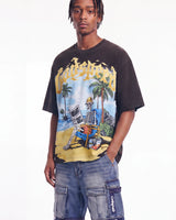 A man wearing a faded black graphic t-shirt with a colorful skeleton on a beach print and the word 'Katrina' in yellow across the chest, paired with blue denim cargo shorts, against a plain white background.