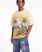 A man wearing a beige Godspeed t-shirt with a colorful graphic of a skeleton on a beach and blue cargo shorts, photographed against a plain white background.