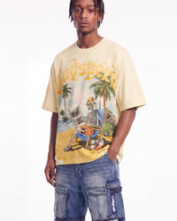 A man wearing a beige Godspeed t-shirt with a colorful graphic of a skeleton on a beach and blue cargo shorts, photographed against a plain white background.