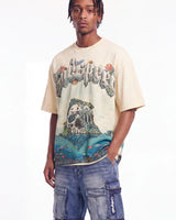 A man wearing a cream-colored graphic t-shirt with a detailed tropical island and skull print, paired with blue denim cargo shorts, against a plain white background.