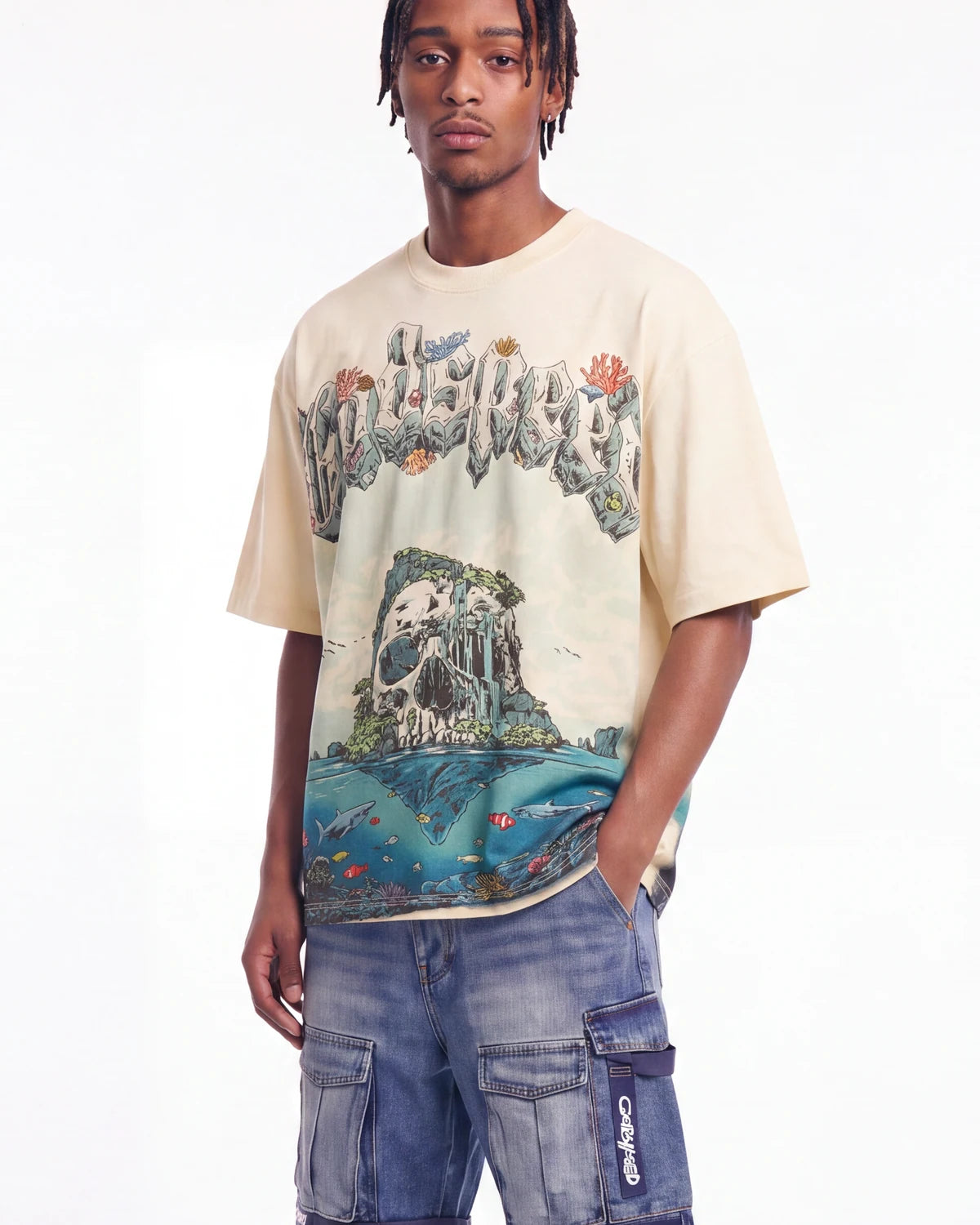 A man wearing a cream-colored graphic t-shirt with a detailed tropical island and skull print, paired with blue denim cargo shorts, against a plain white background.