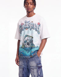 A man wearing a white graphic t-shirt with a colorful 'The Alchemist' design and blue cargo pants, photographed against a plain white background.