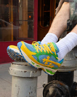Bravest Studios Kross Kountry Runner Brazil, worn on foot propped on a sidewalk bollard showing the yellow laces and green midsole