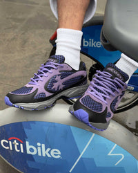 Bravest Studios Kross Kountry Runner Dirty Soda Purple, worn on foot resting on a Citi Bike dock showing the lilac laces and navy mesh