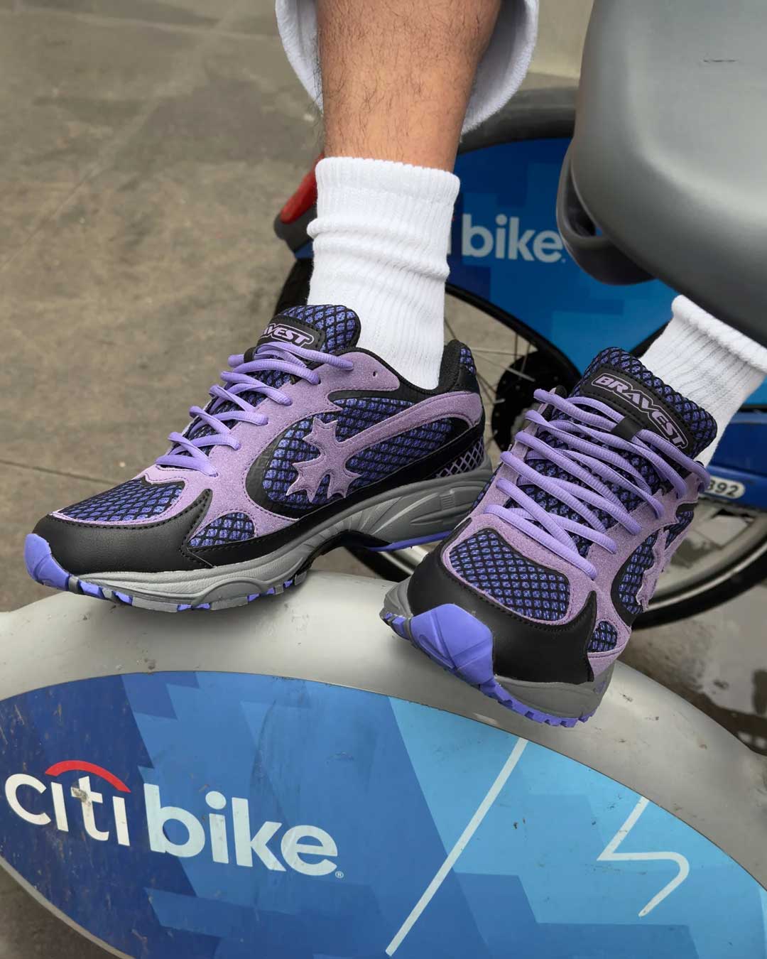 Bravest Studios Kross Kountry Runner Dirty Soda Purple, worn on foot resting on a Citi Bike dock showing the lilac laces and navy mesh