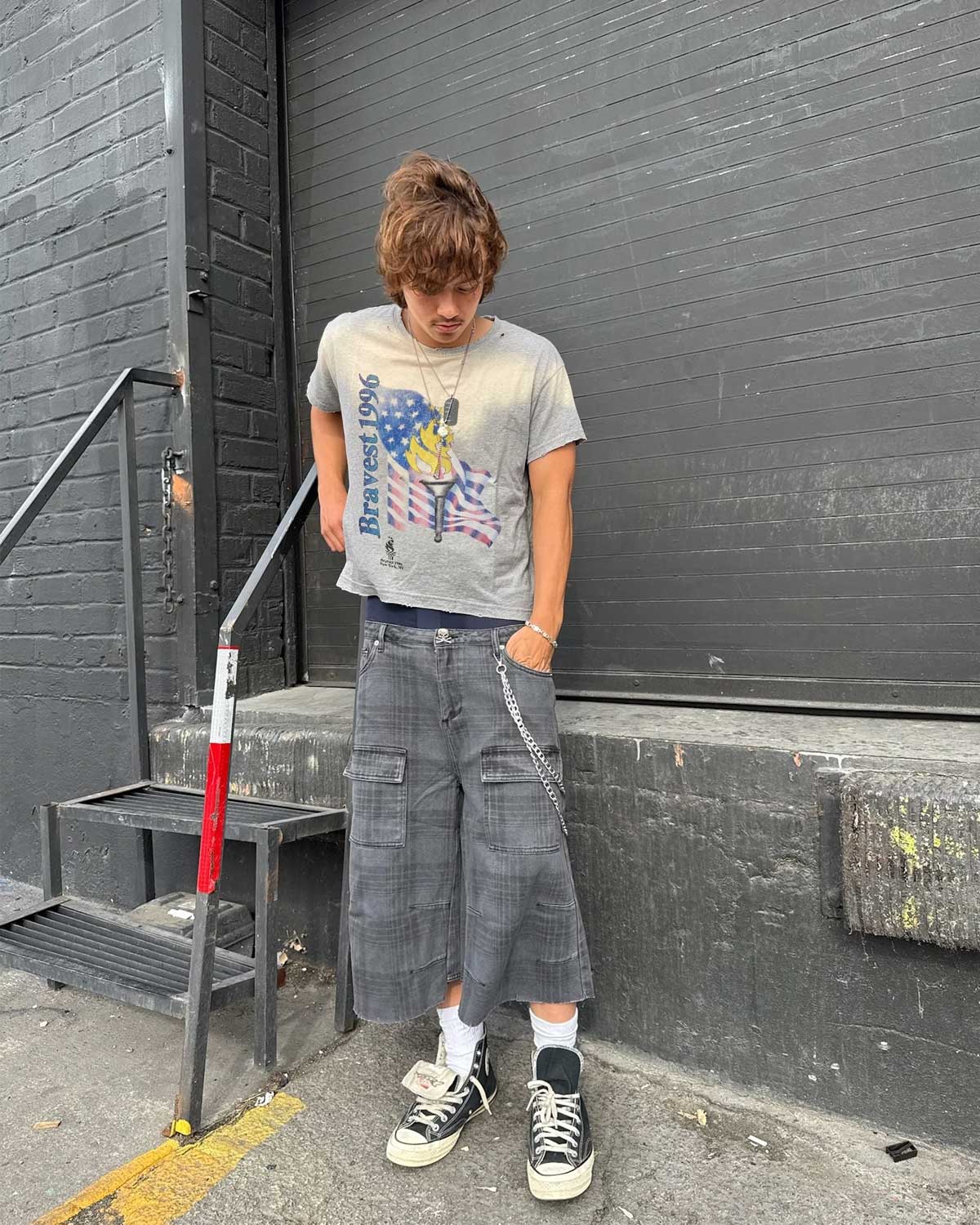 A person standing outdoors in front of a dark gray brick wall and metal steps, wearing a light gray graphic T-shirt, loose dark gray cropped cargo shorts, white socks, and black low-top sneakers.