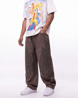 A person wearing loose brown patchwork cargo pants, a white graphic t-shirt, and white sneakers, shown from the chest down against a plain white studio background.