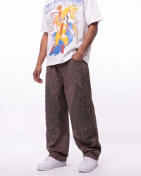 A person wearing loose brown patchwork cargo pants, a white graphic t-shirt, and white sneakers, shown from the chest down against a plain white studio background.