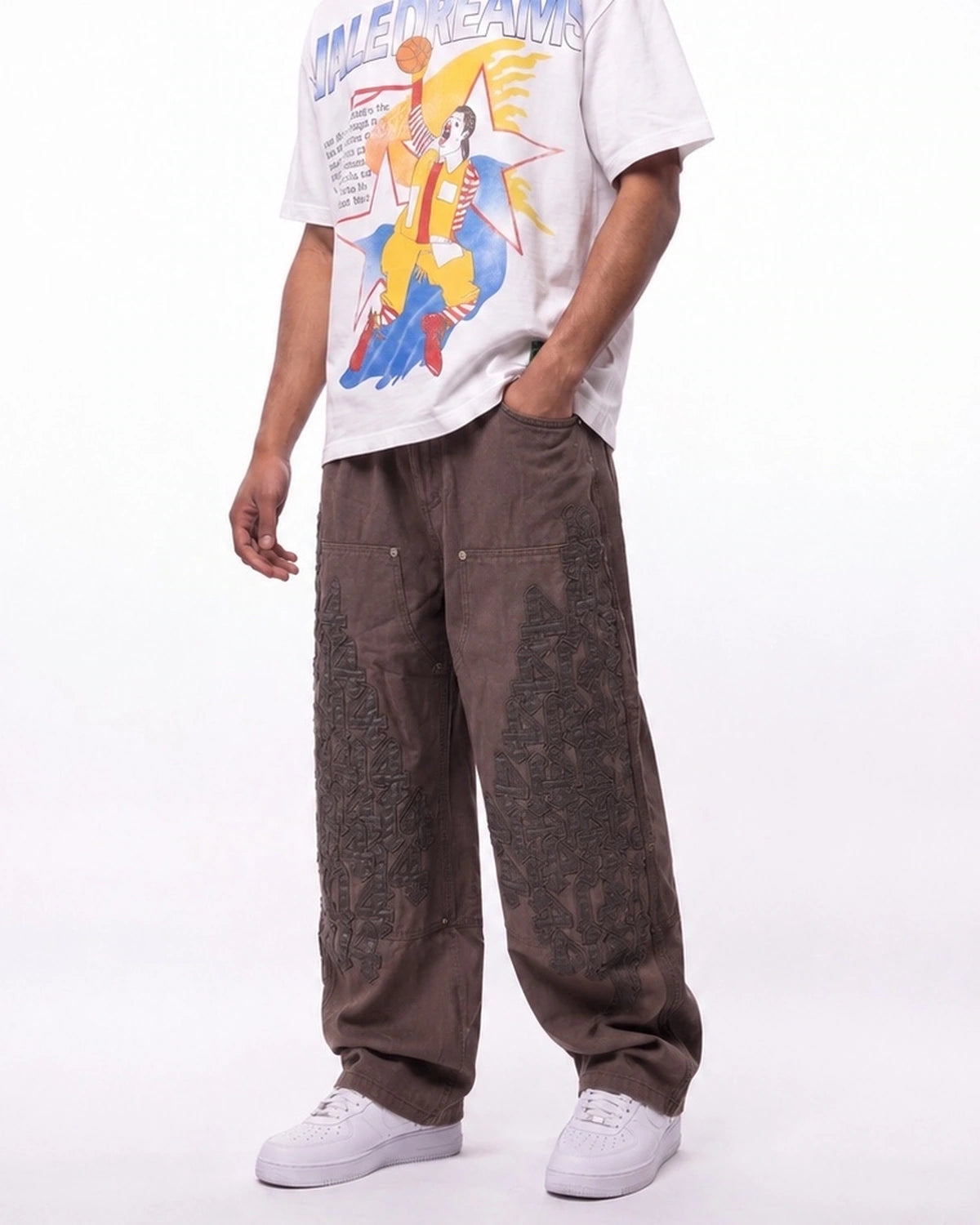 A person wearing loose brown patchwork cargo pants, a white graphic t-shirt, and white sneakers, shown from the chest down against a plain white studio background.