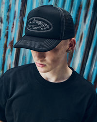 Corteiz Island Outline Trucker Hat black, worn by a model with a black tee, close front view