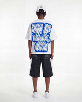 A model shown from the back wearing a white t-shirt with a large blue and white graphic text print reading 'NOT OF MANY ONE', black shorts with white side stripes, a white cap, and white sneakers, set against a plain white studio background.