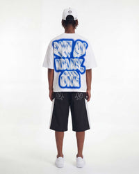 A model shown from the back wearing a white t-shirt with a large blue and white graphic text print reading 'NOT OF MANY ONE', black shorts with white side stripes, a white cap, and white sneakers, set against a plain white studio background.