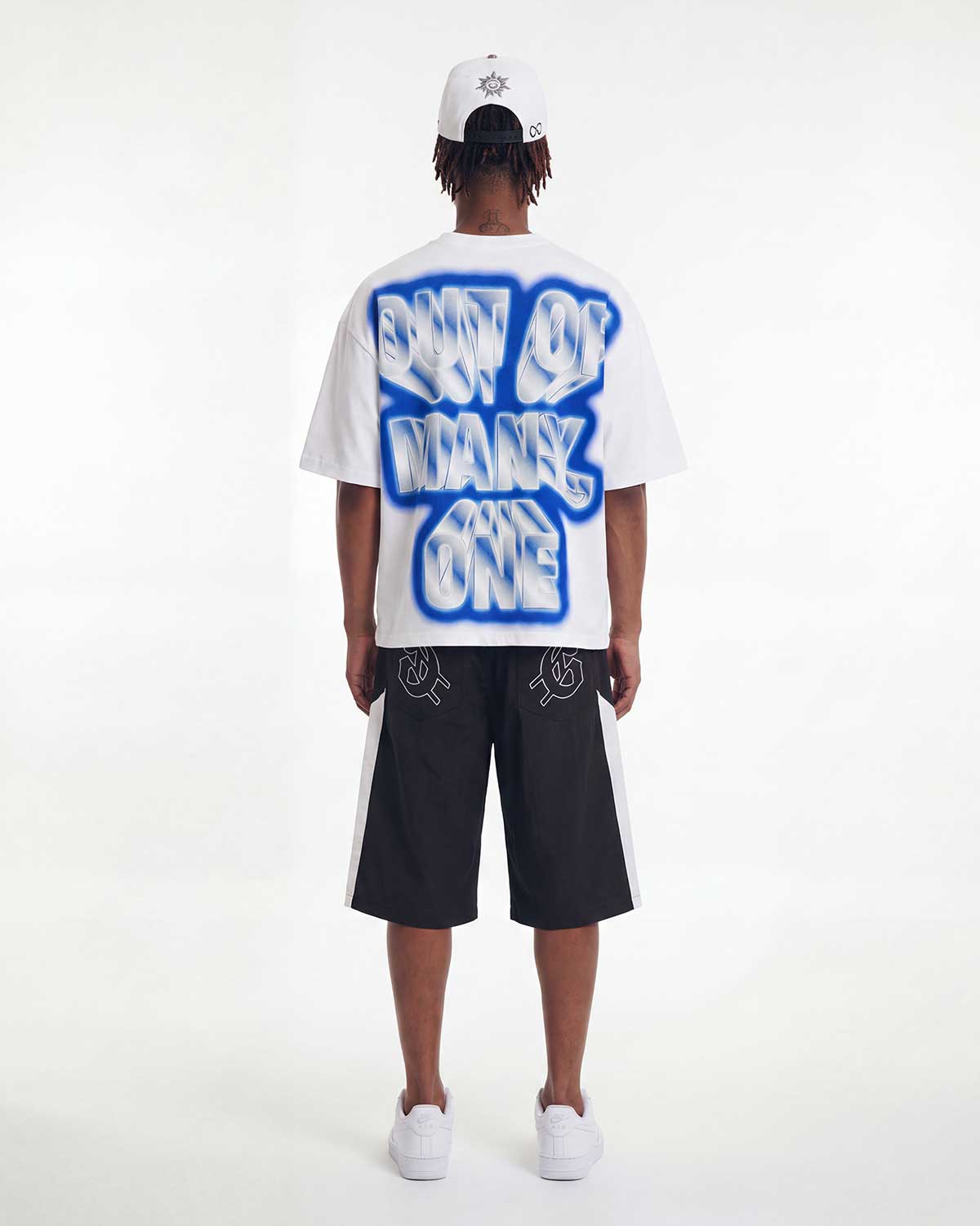 A model shown from the back wearing a white t-shirt with a large blue and white graphic text print reading 'NOT OF MANY ONE', black shorts with white side stripes, a white cap, and white sneakers, set against a plain white studio background.