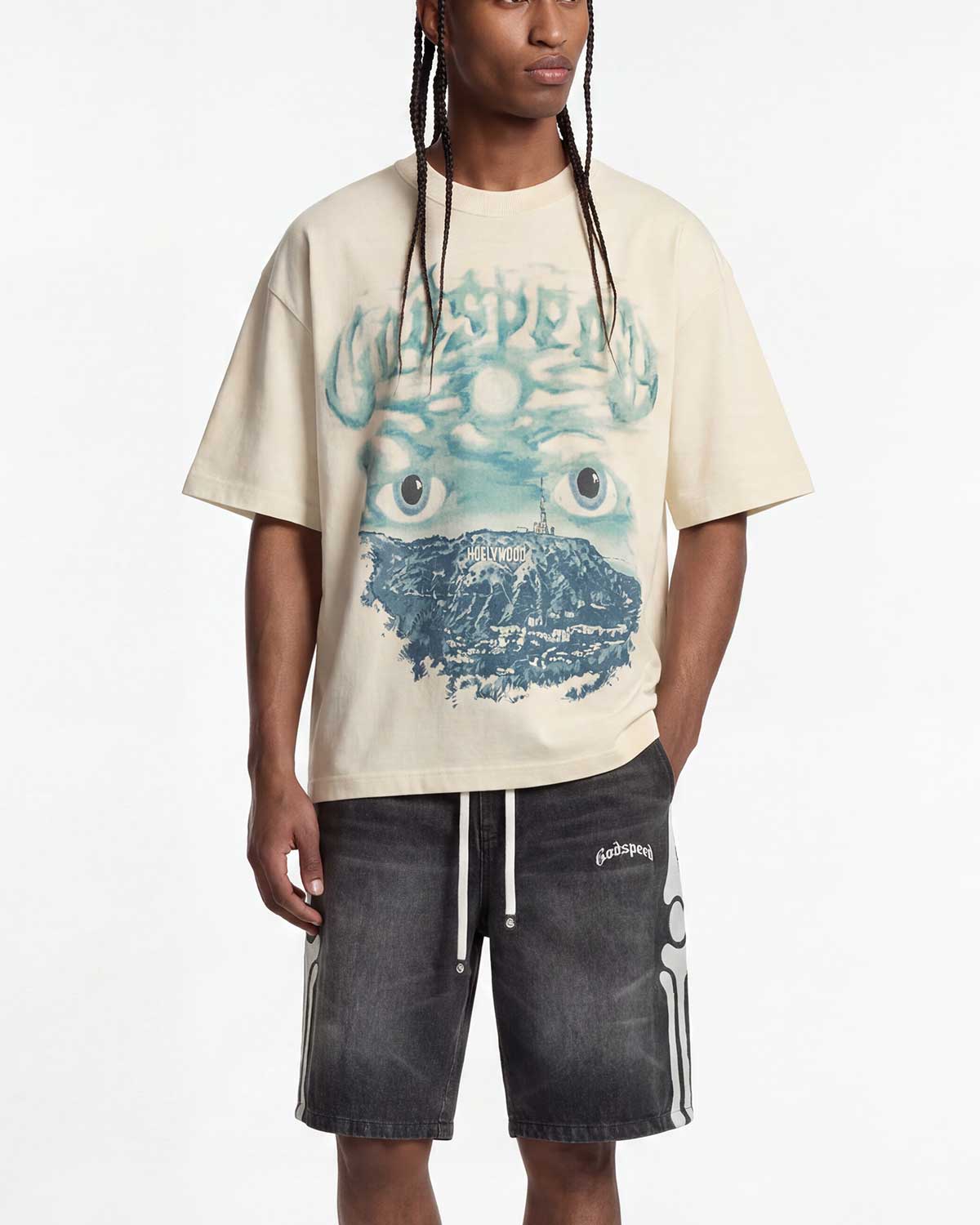 A model wearing a cream-colored t-shirt with a blue and green surreal graphic print of a mountain and eyes, paired with dark gray shorts, against a plain white background.