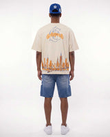A man viewed from behind wearing a cream-colored t-shirt with a large orange and blue graphic print on the back, paired with blue denim shorts and a blue baseball cap.