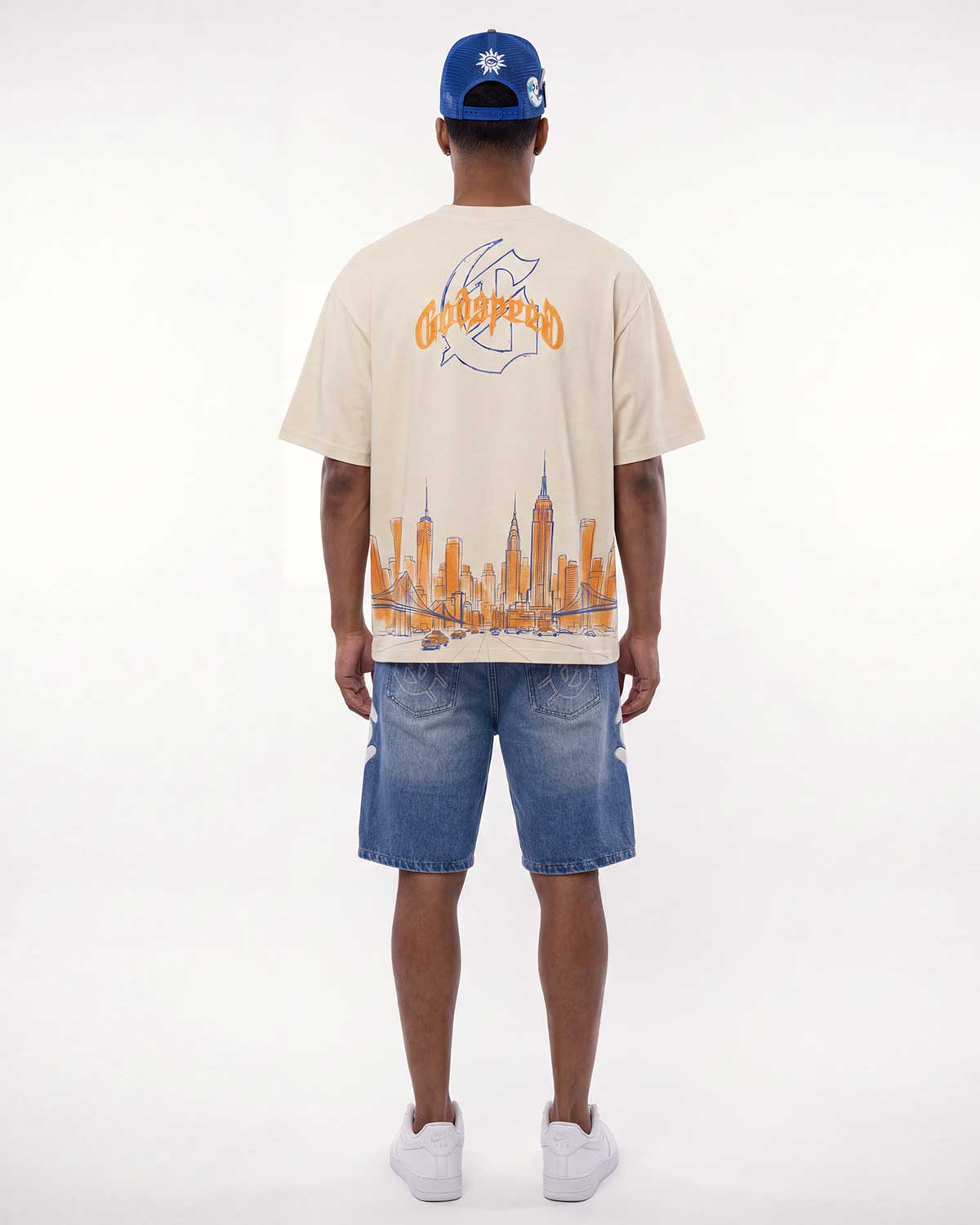 A man viewed from behind wearing a cream-colored t-shirt with a large orange and blue graphic print on the back, paired with blue denim shorts and a blue baseball cap.