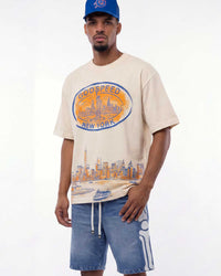 A man wearing a beige Godspeed New York graphic t-shirt with a blue baseball cap and blue denim shorts, standing against a plain white studio background.