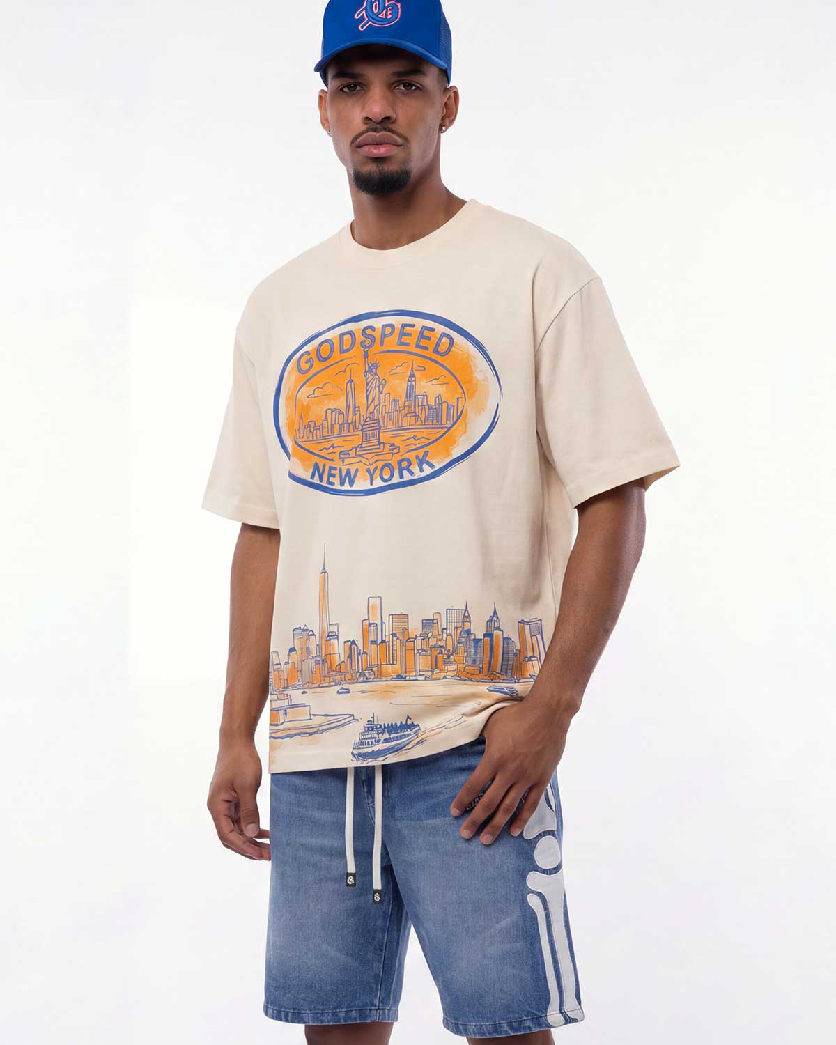 A man wearing a beige Godspeed New York graphic t-shirt with a blue baseball cap and blue denim shorts, standing against a plain white studio background.