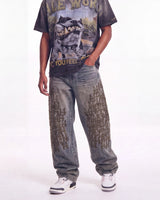 A person modeling oversized faded gray jeans with distressed details and black graphic text patterns, paired with a dark graphic t-shirt and white sneakers, against a plain white studio background.
